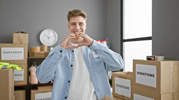 Handsome young caucasian man working hard, confidently volunteering at the heart of community charity center, his smile is infectious as he stands enjoying altruism, forming a lovely heart gesture