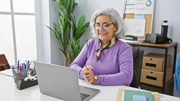 灰色の髪を持つ笑顔の中年の女性は 明るいオフィスで机に座り ラップトップに取り組んでいる — ストック写真