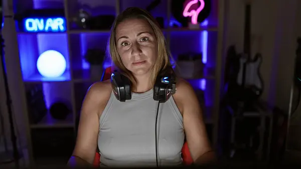 Beautiful, young, blonde female gamer and streamer, earnestly engaged in a nighttime gaming stream, seated in a tech-packed gaming room, geared with headphones and laser-focused on the monitor.