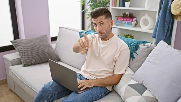 Young focused hispanic man, saying no, using tech-savvy laptop finger gesture at home indoors while sitting alone, serious but concentrated in living room background.
