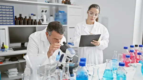Male and female scientists working together in lab, serious concentration on their research, writing on clipboard, using microscope