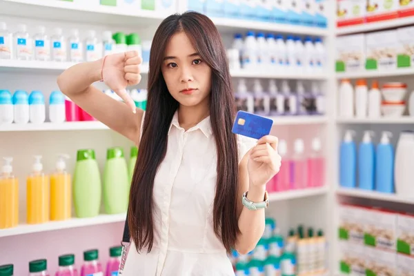 Young chinese woman shopping at pharmacy drugstore holding credit card with angry face, negative sign showing dislike with thumbs down, rejection concept