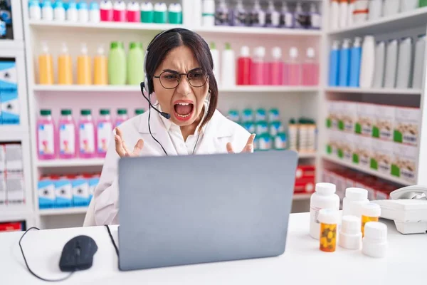 Young arab woman working at pharmacy drugstore using laptop crazy and mad shouting and yelling with aggressive expression and arms raised. frustration concept.