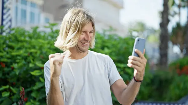 A cheerful young man with long blonde hair celebrating while looking at a smartphone outdoors in a lush garden.