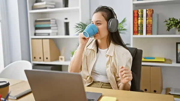 Hard-at-work young hispanic woman, an attractive business pro, indulges in her favorite song while sipping on coffee amidst her elegant office backdrop.