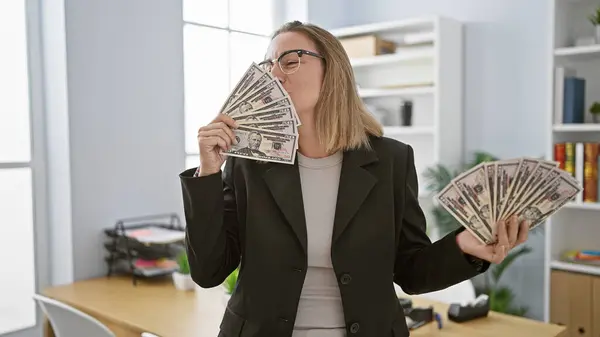 In the office, a beautiful blonde woman kissing dollars, young, confident business executive celebrating success in her workplace