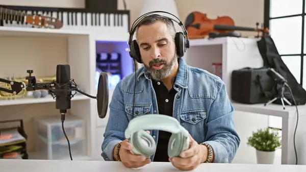 stock image A mature hispanic man with a beard and grey hair holds headphones inside a music studio with microphones and instruments.