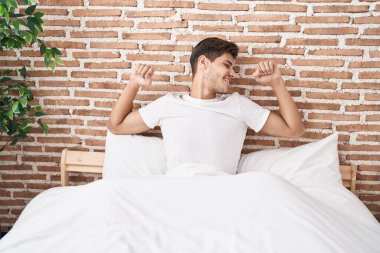 Young hispanic man waking up stretching arms at bedroom
