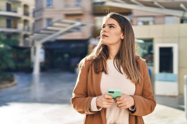 Genç, güzel, İspanyol bir kadın sokakta akıllı telefon kullanarak gülümsüyor.