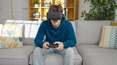 Evde video oyunu oynayan İspanyol genç adam joystick ve sanal gerçeklik gözlüğü kullanıyor.