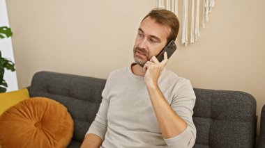 Orta yaşlı, sakallı bir adam modern bir oturma odasında otururken telefonla konuşuyor..