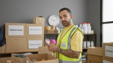 Sakallı, dövmeli, gönüllü yelek giyen İspanyol bir adam bir depoda bağış kutularıyla duruyor..