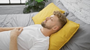 Genç sakallı adam, sarı yastıklı ve gri yataklı modern bir yatak odasında huzur içinde uyuyor..