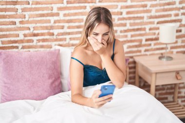 Young blonde woman using smartphone with surprise expression at bedroom