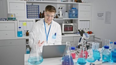 Yakışıklı beyaz erkek-bilim adamı. Laboratuvarda sürükleyici bir video görüşmesi yaparken gülümsüyor. Tıp araştırmaları, teknoloji ve test tüpleri arasında..