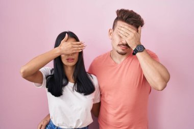 Young hispanic couple standing over pink background covering eyes with hand, looking serious and sad. sightless, hiding and rejection concept 