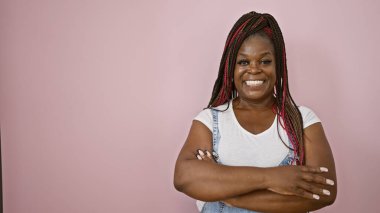 Kendinden emin Afro-Amerikan kadını, neşeli bir şekilde gülümsüyor, kollarını kavuşturup, izole pembe bir arka planda rahat bir jest yapıyor..