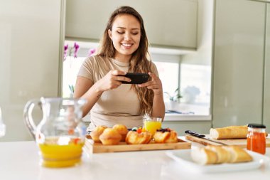 Young beautiful hispanic woman make photo by smartphone to breakfast at the kitchen