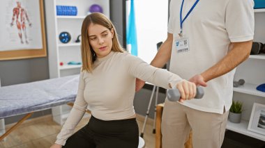 Bir kadın hasta, rehabilitasyon kliniğinde erkek bir fizyoterapistin yardımıyla bir halterle kol egzersizi yapıyor..