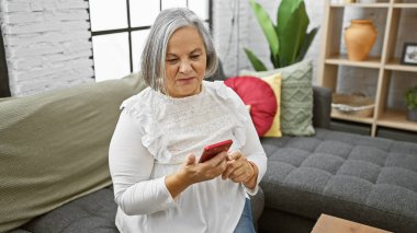 Beyaz saçlı, rahat, modern bir oturma odasında akıllı telefon kullanan son sınıf öğrencisi kadın.