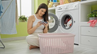 Elbiselerini yıkayan genç, İspanyol bir kadın çamaşır odasında akıllı telefonla oyun oynuyor.