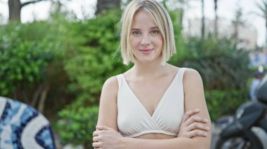 Kollarını kavuşturmuş genç sarışın kadın parkta gülümsüyor.