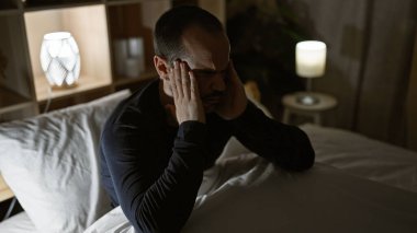 Kel, kel, sakallı bir adam gecenin karanlığında karanlık bir yatak odasında oturuyor. Yüzünde baş ağrısı ya da endişe ifadesi var..