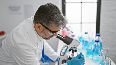 Çekici gri saçlı İspanyol bir adam, kendini adamış bir bilim adamı, laboratuarda tıbbi bir deney üzerinde çalışıyor, güvenlik eldivenleri takılı, mikroskoptan dikkatle bakıyor,.