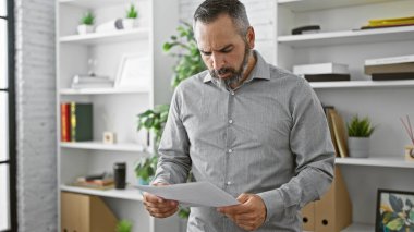 Sakallı, kır saçlı, modern bir ofiste doküman okuyan, işinde üst düzey bir profesyoneli canlandıran olgun İspanyol bir adam..