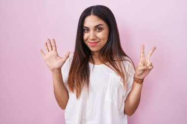 Pembe arka planda duran genç Arap kadın yedinci parmaklarını gösterip işaret ederken kendinden emin ve mutlu bir şekilde gülümsüyor.. 