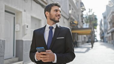 Yakışıklı İspanyol takım elbiseli adam güneşli şehir caddesinde akıllı telefon kullanıyor..