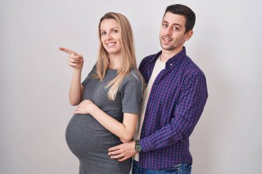 Genç bir çift beyaz arka planda duran bir bebek bekliyor. Arka tarafı işaret ediyor. Başparmakları havada, kendinden emin bir şekilde. 