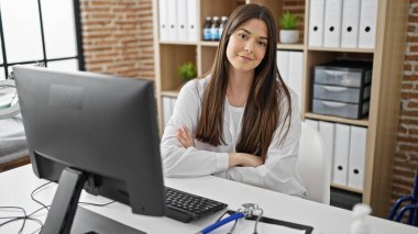 Kollarını kavuşturup klinikte gülümseyen genç ve güzel İspanyol kadın doktor.