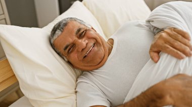 Gri saçlı, gülümseyen orta yaşlı bir adam yatak odasında rahatlık ve mutluluk tasvir ediyor..