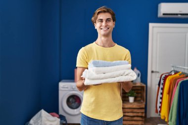 Young caucasian man smiling confident holding folded towels at laundry room