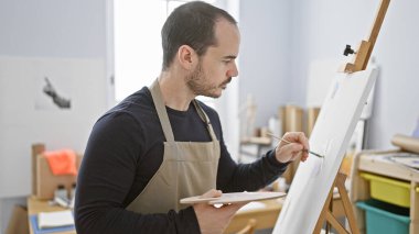Kel, sakallı, resim stüdyosunda tuvale resim çizen, önlük takan ve dikkatle odaklanan bir İspanyol..