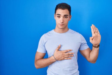 Young hispanic man standing over blue background swearing with hand on chest and open palm, making a loyalty promise oath 