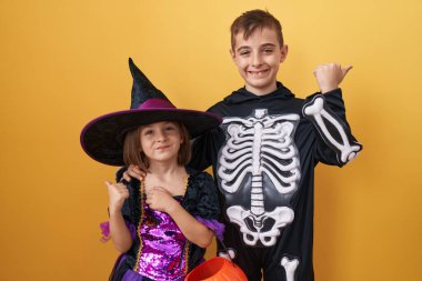 Cadılar Bayramı kostümleri içinde neşeli erkek ve kız kardeşler başparmakları havada, sarı izole edilmiş arka planda ağzı açık gülümsemeleri var.