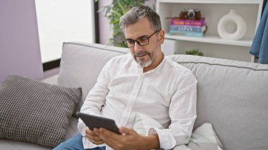 Kendine güveni tam, beyaz saçlı bir adam dokunmatik ped kullanırken rahat bir şekilde kanepede oturuyor ve evde dinleniyor, evde pozitif yaşam tarzı sergiliyor..