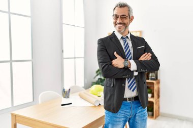 Orta yaşlı, kır saçlı mimar ofiste kollarını kavuşturmuş duruyor.