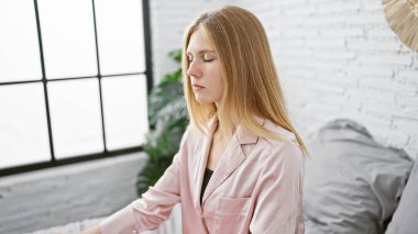 Pembe gömlekli sarışın kadın, parlak beyaz, modern bir yatak odasında, sakin ve düşünceli görünüyor.