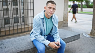 Şehir ortamında akıllı telefon kullanan İspanyol bir adam bankta oturuyor.