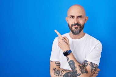 Mavi arka planda dikilen İspanyol dövmeli bir adam. El parmağı yanda, ciddi ve sakin bir yüz ifadesi sergiliyor. 