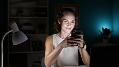 Loş bir ofis odasında akıllı telefon kullanarak gülümseyen genç bir kadın..