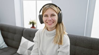 Gülümseyen beyaz kadın, sarı saçlı ve mavi gözlü modern bir oturma odasında kulaklık takıyor.