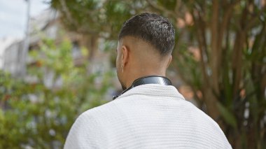 Modern saç stili ve küpe takan, çevresi yeşilliklerle çevrili şık genç bir adamın arka planı..