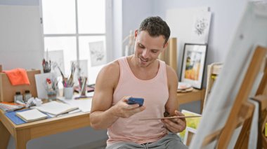 Sanat stüdyosunda resim ve sanat malzemeleriyle çevrili akıllı telefon kullanan yakışıklı bir adam.