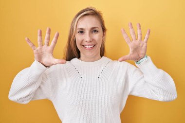 Beyaz süveter giyen, sarı arka planda beyaz bir kadın on numarayı gösteriyor ve gülümsüyor, kendinden emin ve mutlu.. 