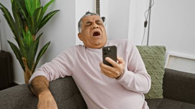 Mature man yawning with smartphone in a cozy living room, expressing fatigue or boredom. clipart