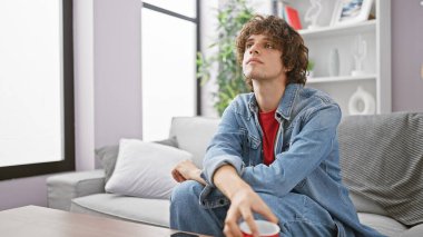 Modern bir oturma odasında uzanan kıvırcık saçlı yakışıklı genç adam, düşünceli görünüyor..